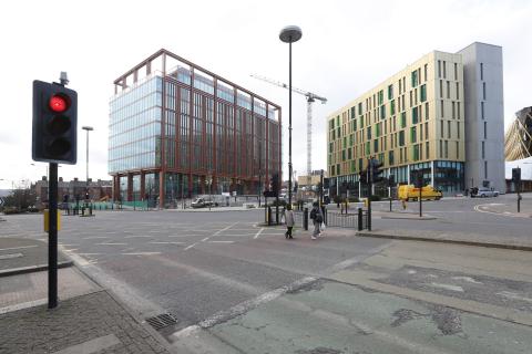 A photo of a junction with traffic lights in the foreground, two people crossing the road and buildings a the other side of the junction.